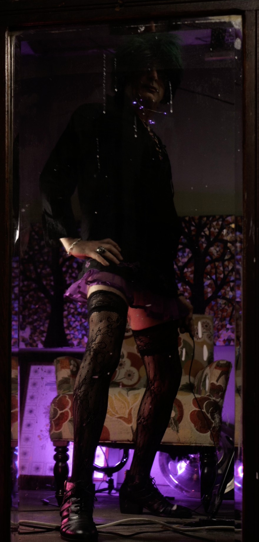 a goth woman sitting in an opulent chair in a moody room, her left left kicks straight up, her short skirt has purple ruffles... magenta and blue lighting